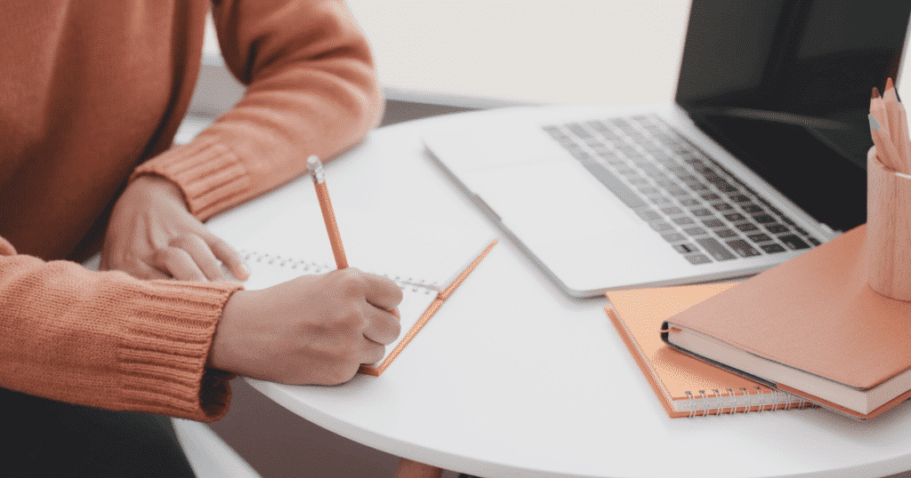 A person writing on a notebook with a laptop and extra notebook on the side