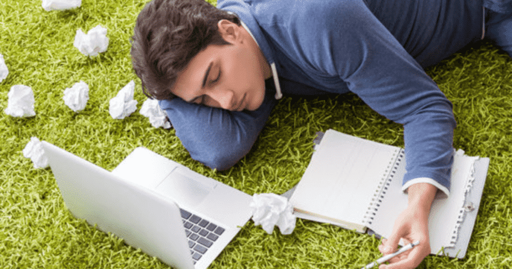 A person lying down on the grass besides a labtop and folded pieces of paper